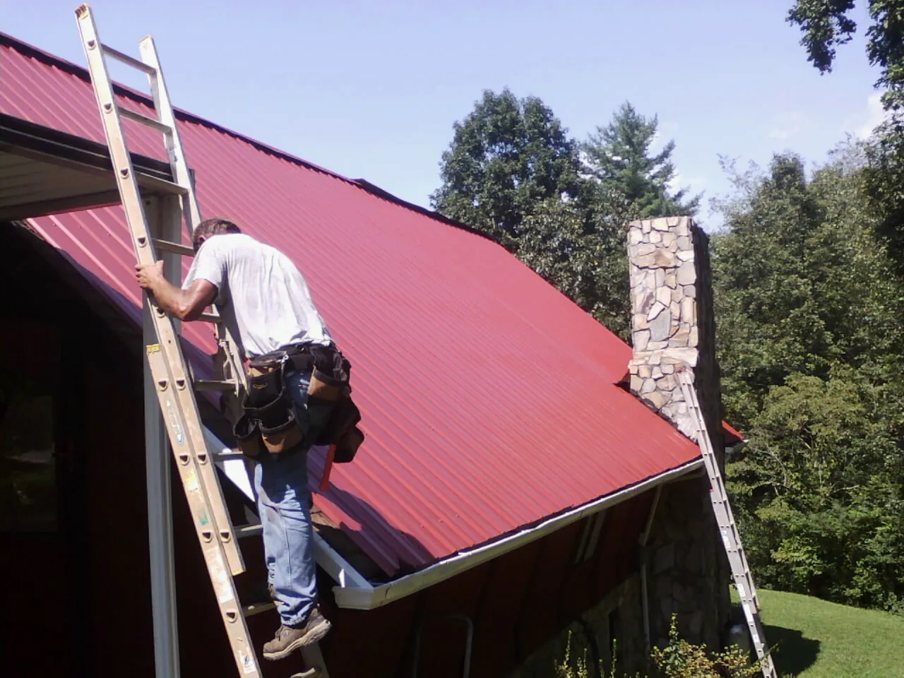 Roofer on a ladder beside a red metal roof and stone chimney — Soffit & Fascia Repair by Roofing Brothers in Dunfermline